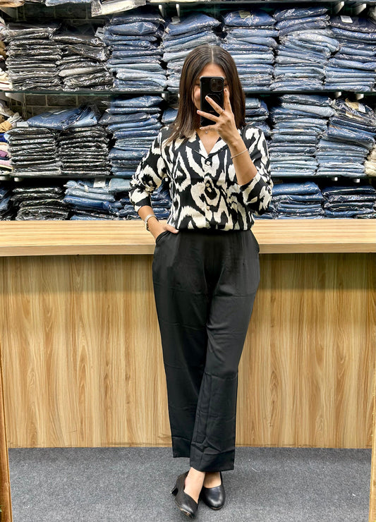 Woman wearing black high-waisted pants and black white printed top posing in front of folded denim stacks
