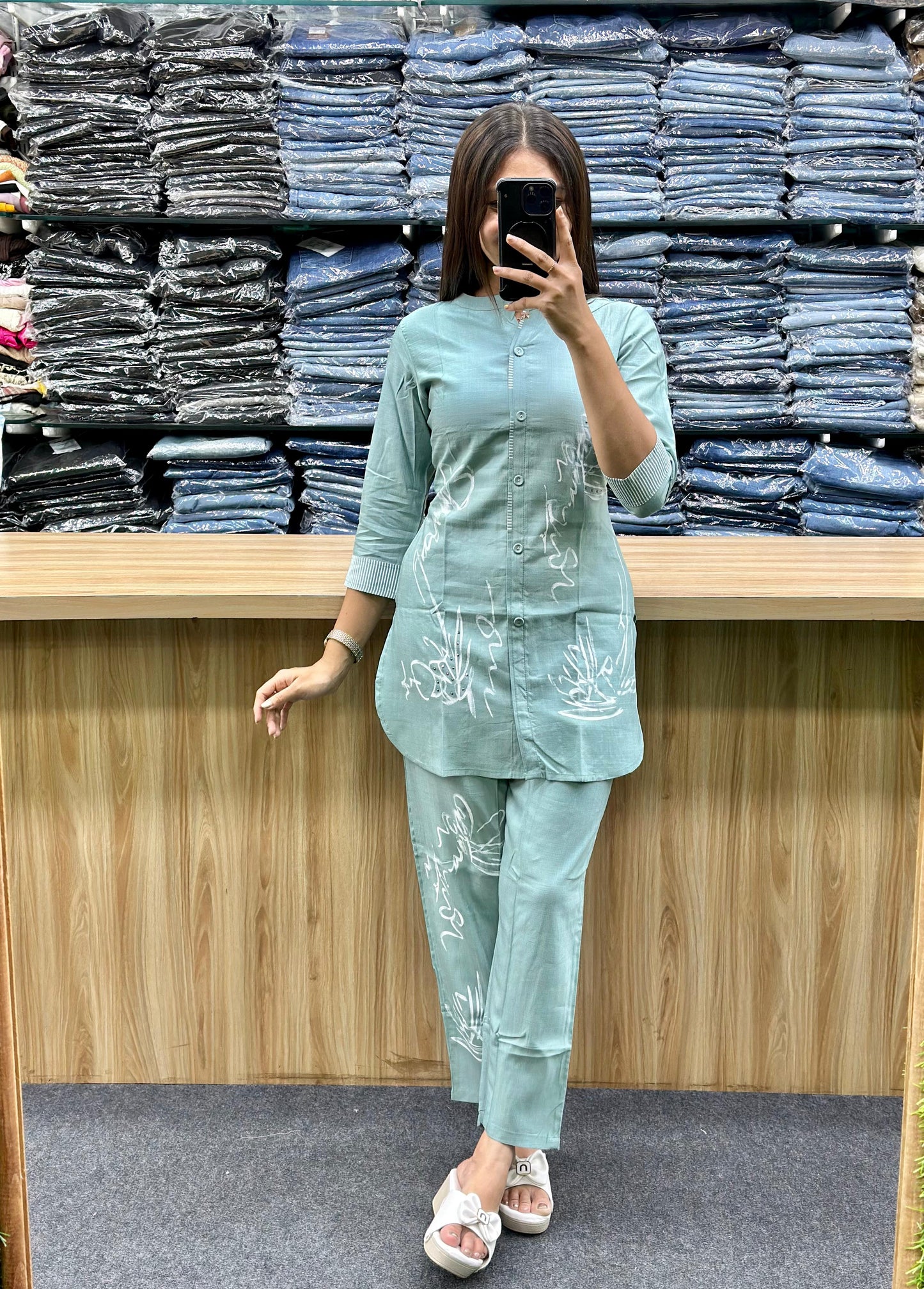Woman wearing light blue cotton loungewear set with subtle white embroidery standing in front of stacked denim jeans