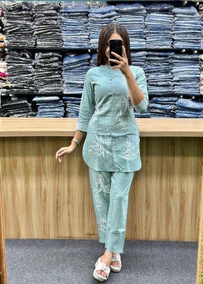 Woman wearing light blue cotton loungewear set with subtle white embroidery standing in front of stacked denim jeans