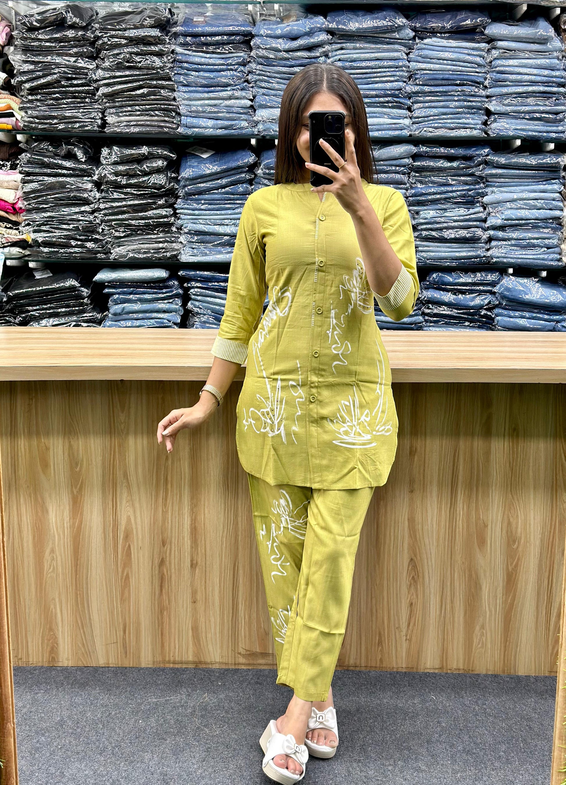 Woman wearing yellow cotton loungewear set with white floral designs standing in front of folded denim stacks