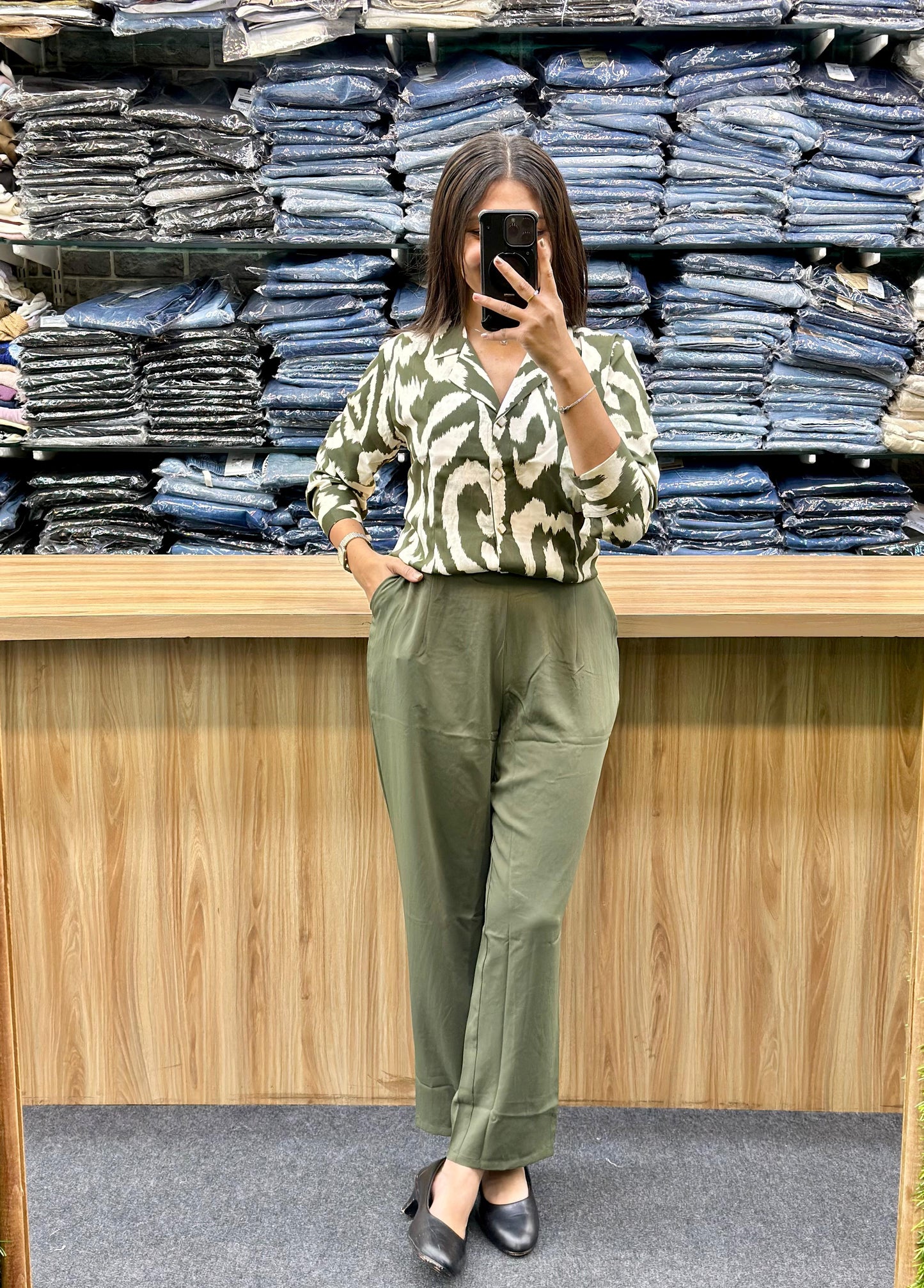 Woman wearing olive green pants and patterned green blouse standing in front of stacked denim jeans at Vinayak Fashion store
