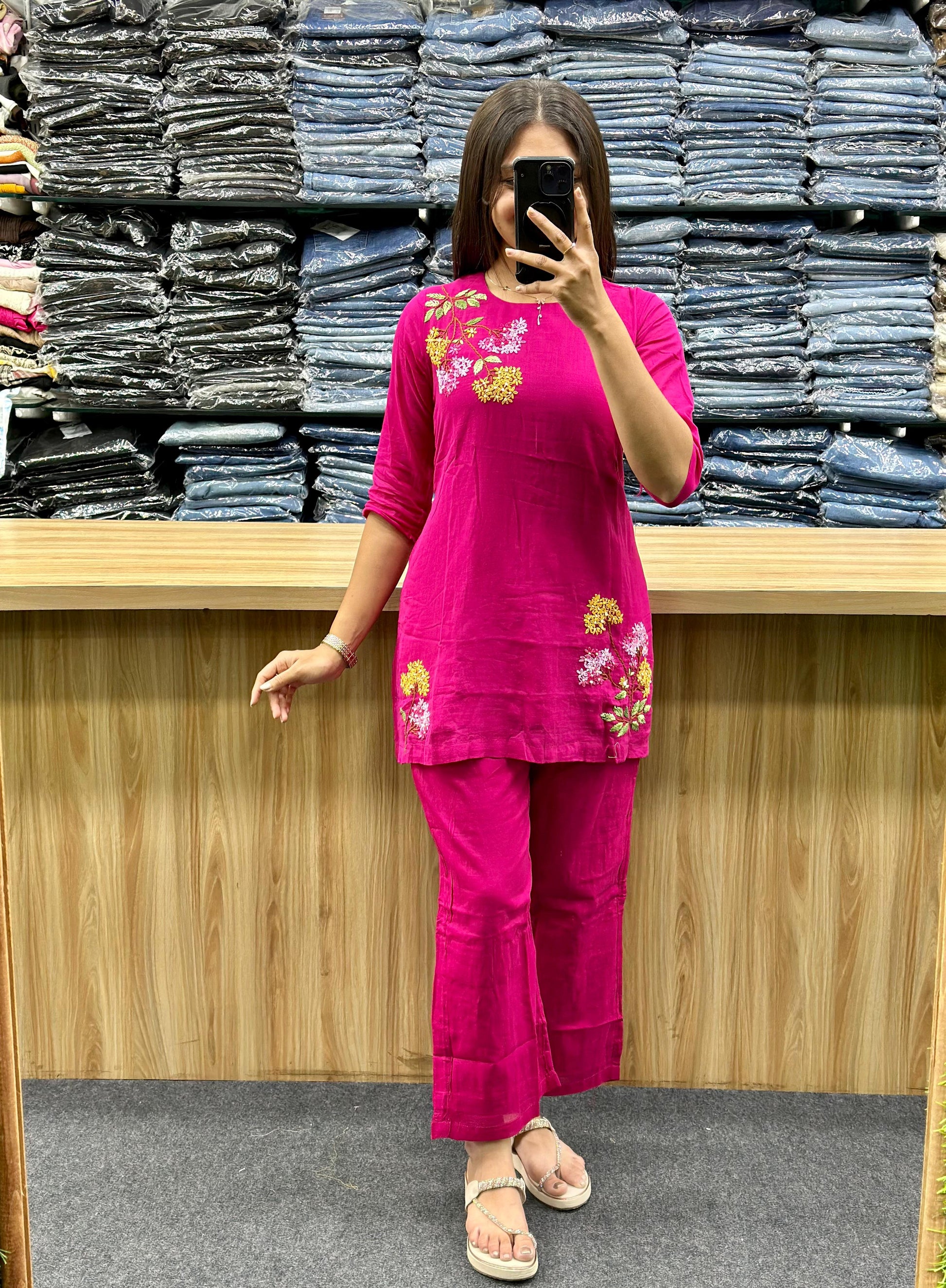 Woman wearing pink cotton co ord set with floral embroidery standing in front of stacked denim clothes