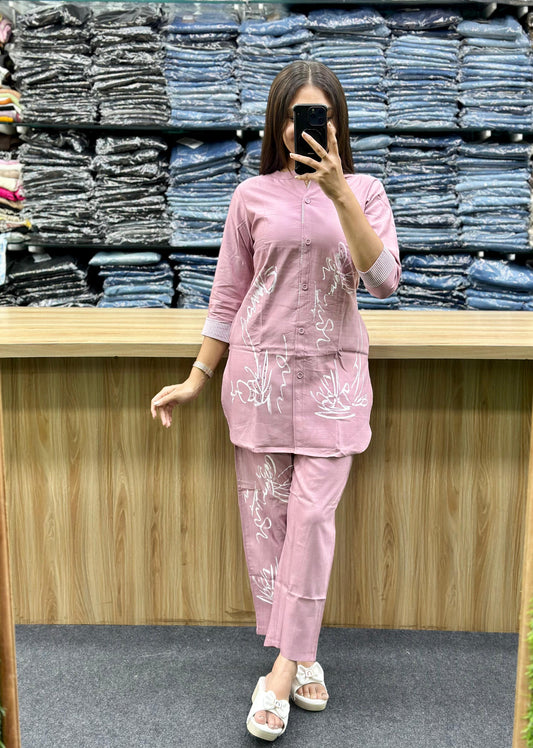 Woman wearing a pink cotton loungewear set with white floral prints standing in front of folded jeans